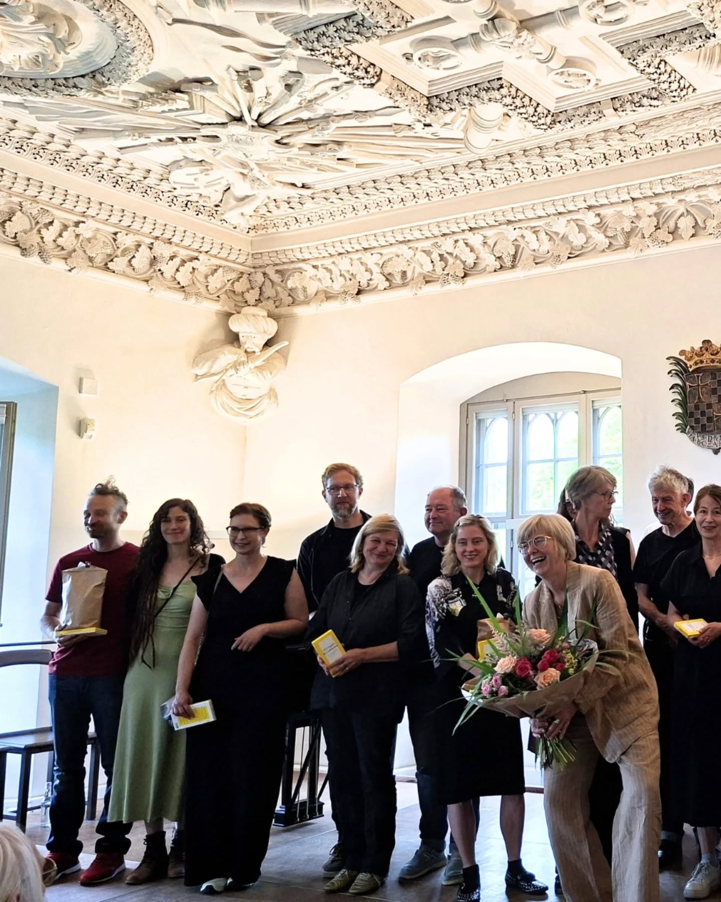 Eine Gruppe von Menschen steht in einem Raum mit einer kunstvollen Decke. Eine Person hält einen Blumenstrauß.