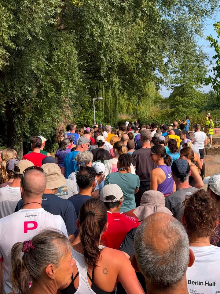 Eine große Menschenmenge steht im Freien unter Bäumen und blickt in die Ferne. Viele tragen Sportkleidung und Startnummern, wobei auf einem weißen Shirt im Vordergrund der Text "Berlin Half" zu sehen ist. (KI-Alternativtext)