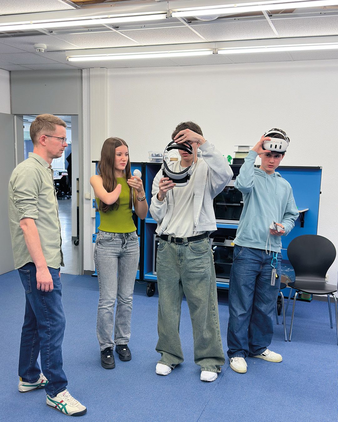 Dieser Alternativtext wurde KI-generiert: Eine Gruppe von Personen steht in einem Raum. Eine Person hält ein Virtual Reality Headset vor dem Gesicht, während andere zusehen und etwas in den Händen halten.