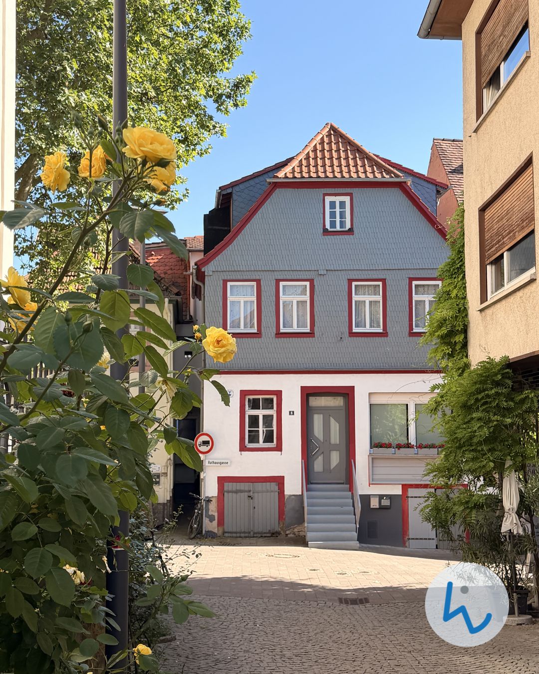 Graues Fachwerkhaus mit roten Fensterrahmen und grauer Tür, umgeben von gelben Rosen und grünen Pflanzen unter blauem Himmel. (KI-Alternativtext)