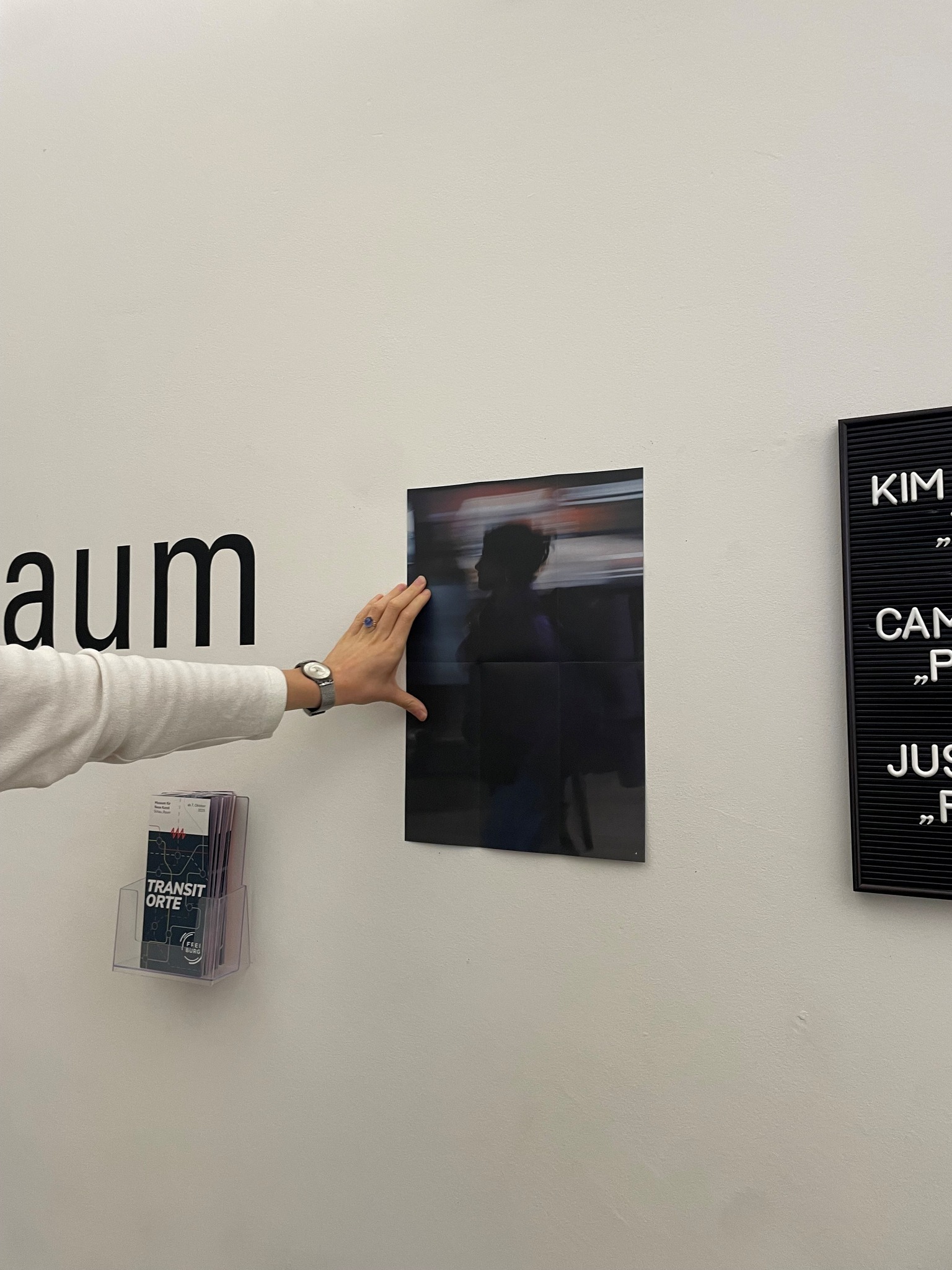Dieser Alternativtext wurde KI-generiert: Ein Schwarz-Weiß-Foto ist an einer weißen Wand aufgehängt und wird von einer Hand berührt. Neben dem Foto befindet sich ein Informationsblatt und ein schwarzes Schild mit Text.