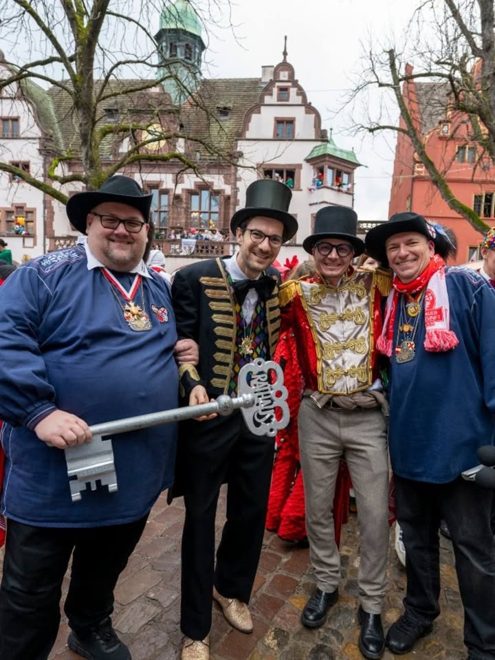 Dieser Alternativtext wurde KI-generiert: Farbfoto zeigt mehrere Personen in festlicher Kleidung, darunter Hüte und Medaillen, vor einem Gebäude mit historischer Architektur. Im Vordergrund hält eine Person einen großen Schlüssel.