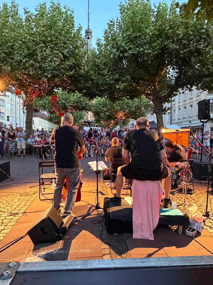 Eine Band mit vier Musikern spielt auf einer offenen Bühne im Freien, wobei die Musiker von hinten zu sehen sind. Im Vordergrund stehen Lautsprecher und Mikrofonständer, während im Hintergrund ein Publikum unter Bäumen mit Lichterketten zuschaut und in der Ferne ein hoher Turm sichtbar ist. (KI-Alternativtext)