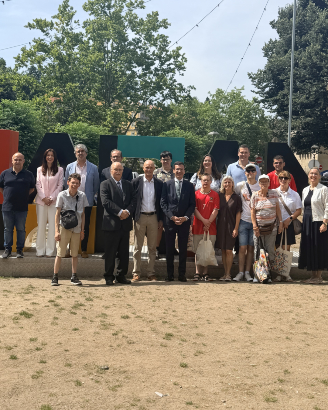 Eine große Gruppe von etwa zwanzig Personen steht im Freien vor einer bunten, geometrischen Wandstruktur für ein Gruppenfoto. Die Kleidung variiert von formellen Anzügen bei einigen Personen in der Mitte bis hin zu Freizeitkleidung wie T-Shirts und Shorts bei den meisten anderen. Im Hintergrund sind grüne Bäume und ein sonniger Himmel zu sehen. (KI-Alternativtext)