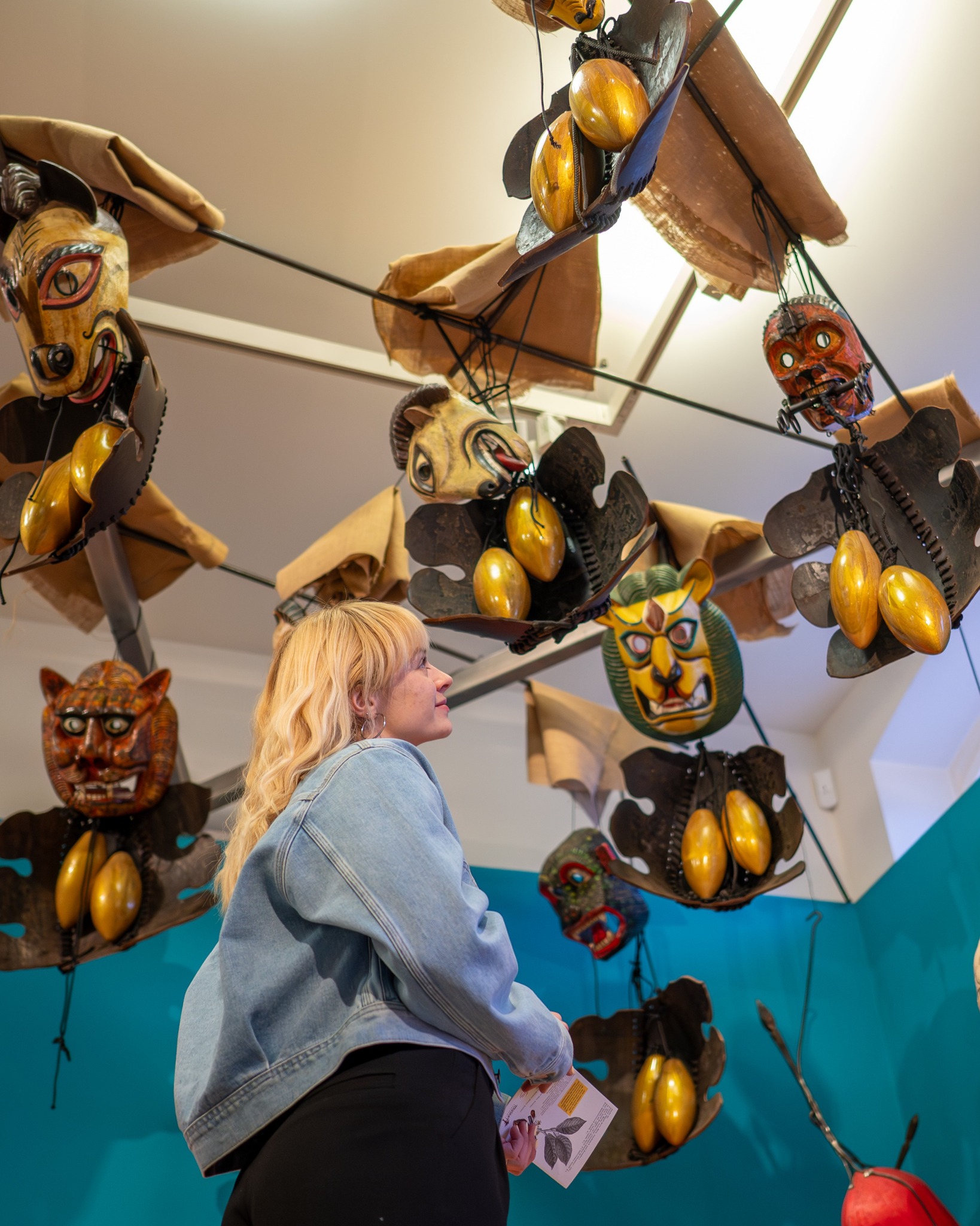 Eine Person mit blonden Haaren und einer hellblauen Jeansjacke blickt nach oben auf eine hängende Kunstinstallation, die aus verschiedenen Masken, goldenen, eiförmigen Objekten und braunen Stoffelementen besteht. Die Person hält eine kleine Informationskarte in der Hand und steht vor einer türkisfarbenen Wand. (KI-Alternativtext)