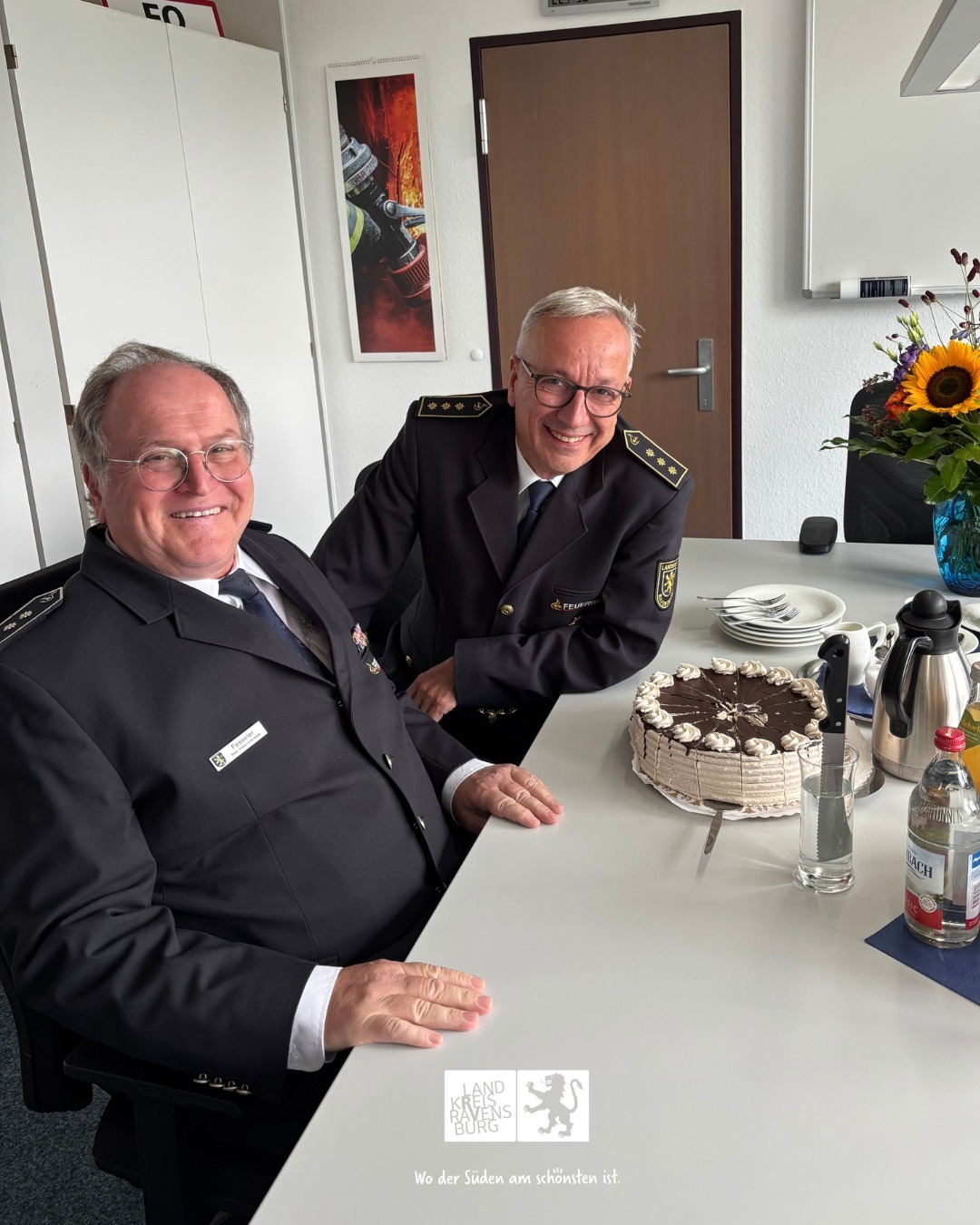 Zwei Personen in Uniform sitzen an einem Tisch mit Kuchen und Getränken. Auf dem Tisch steht ein Blumenstrauß.