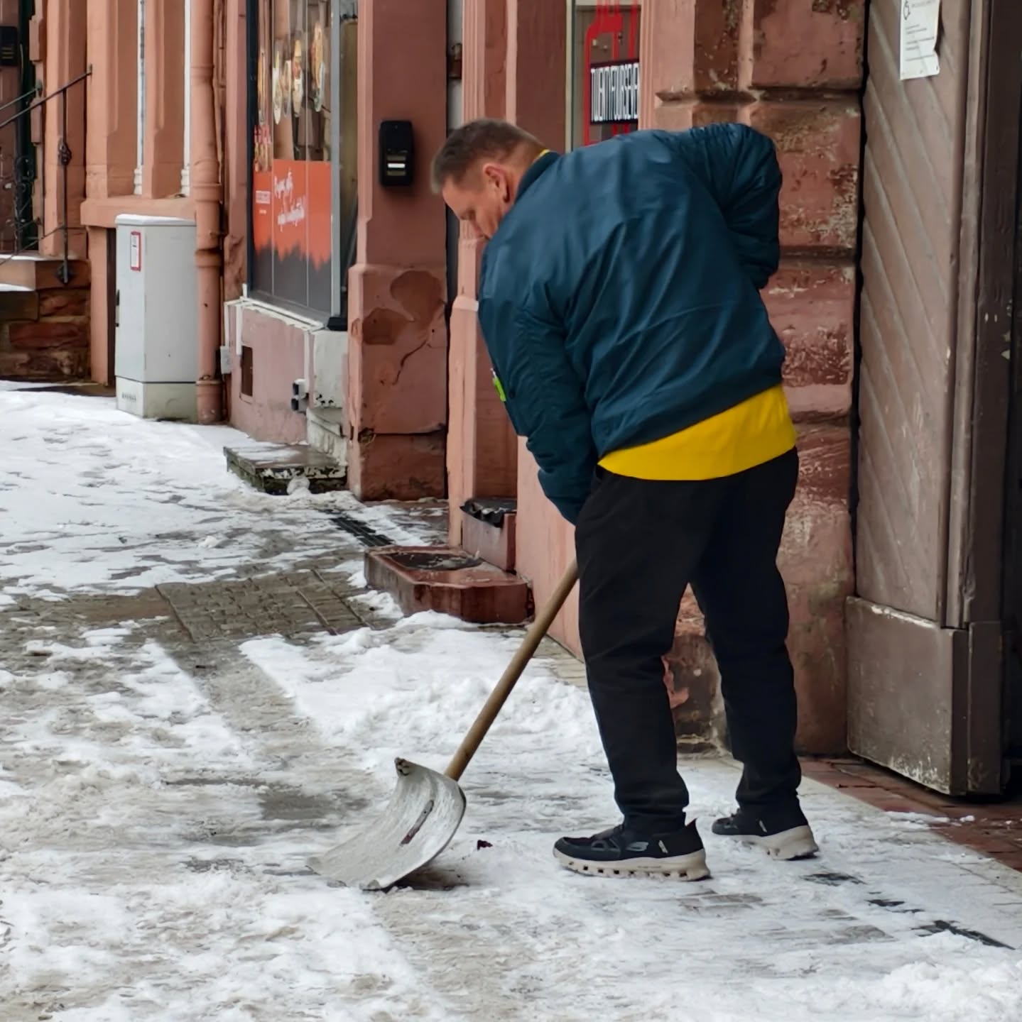 Dieser Alternativtext wurde KI-generiert: Eine Person schaufelt Schnee von einem Gehweg vor einem Gebäude. Das Bild ist farbig und zeigt eine winterliche Szene.