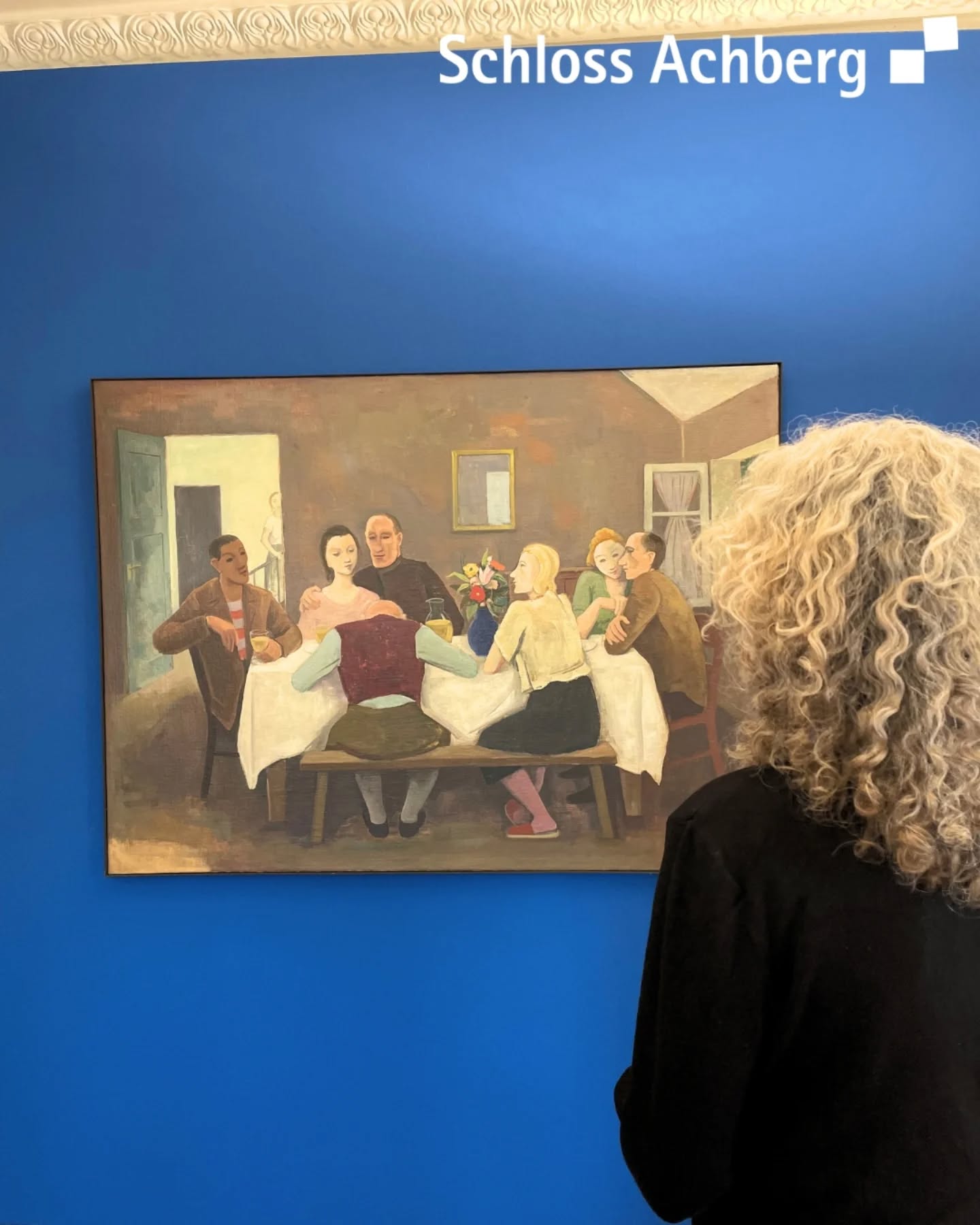 Dieser Alternativtext wurde KI-generiert: Ein Gemälde zeigt eine Gruppe von Personen bei einer Mahlzeit. Im Vordergrund ist ein Teil einer Person zu sehen, die das Bild betrachtet.