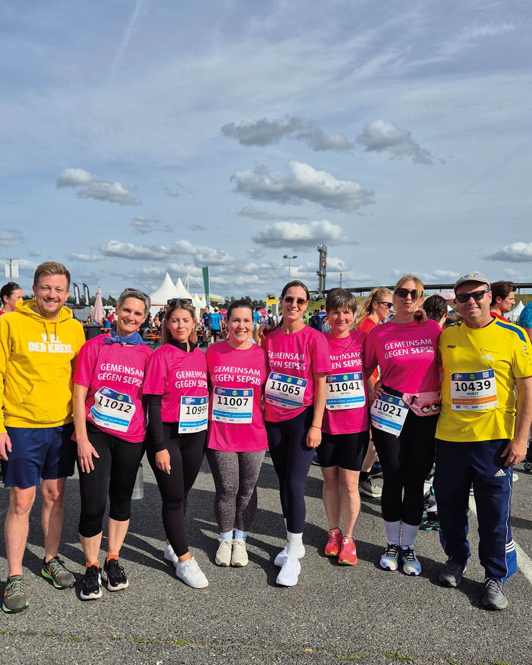 Gruppe von Läuferinnen und Läufern in pinken und gelben Shirts mit Startnummern, die für „Gemeinsam gegen Sepsis“ werben, stehen im Freien vor einem bewölkten Himmel. (KI-Alternativtext)