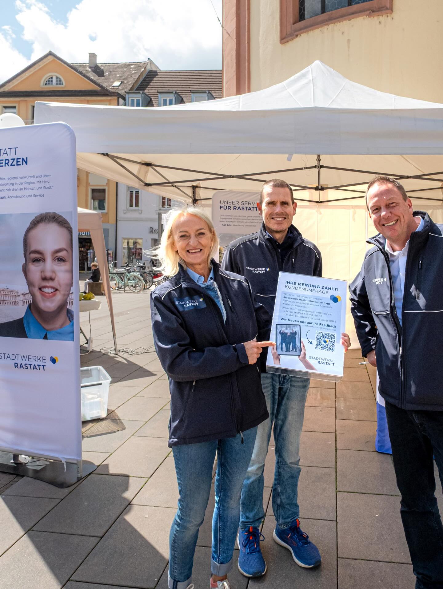 Dieser Alternativtext wurde KI-generiert: Mehrere Personen stehen vor Bannern und einem Stand im Freien. Sie halten ein Schild mit einem QR-Code und lächeln in die Kamera.