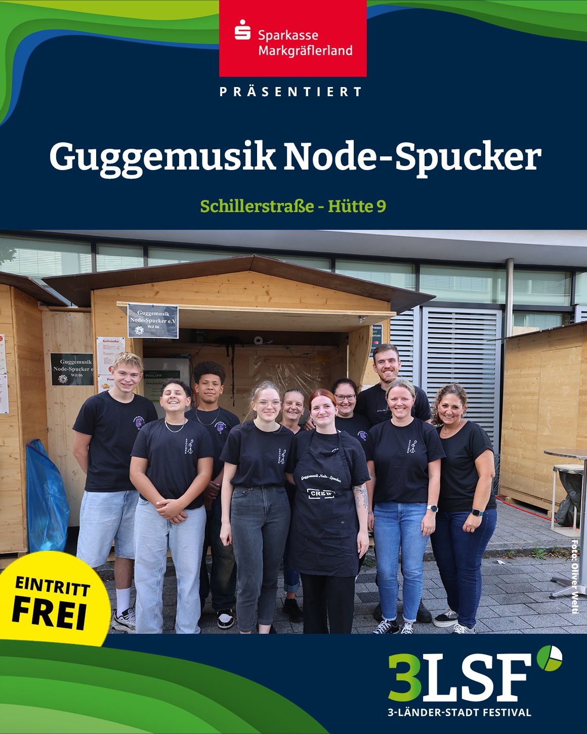 Gruppe von neun Personen in schwarzen T-Shirts vor einem Holzstand mit Schild „Guggemusik Node-Spucker e.V.“; oben Werbung für Sparkasse Markgräflerland und das 3- Länder-Stadt Festival, unten gelber Kreis mit „Eintritt frei“. (KI-Alternativtext)