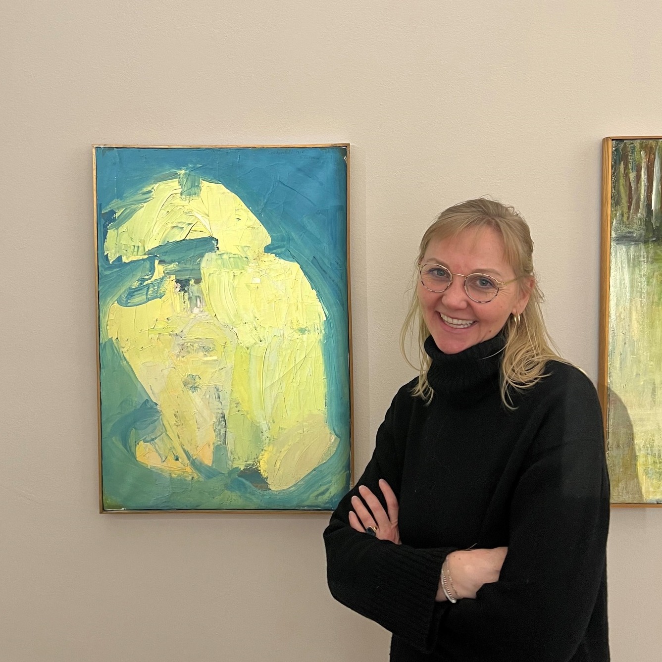 Dieser Alternativtext wurde KI-generiert: Eine Person steht mit verschränkten Armen vor einer hellen Wand. Im Hintergrund hängen zwei farbige Gemälde.