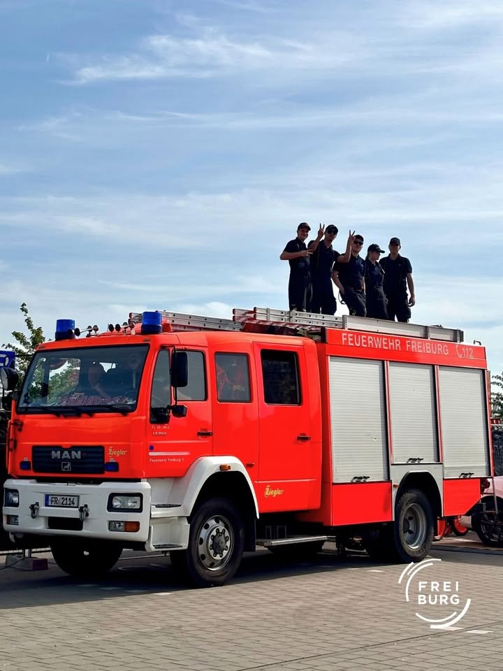 Rotes Feuerwehrfahrzeug der Feuerwehr Freiburg mit der Aufschrift „112“ und mehreren Personen auf dem Dach, die winken. Das Fahrzeug steht auf einem gepflasterten Platz unter blauem Himmel. (KI-Alternativtext)