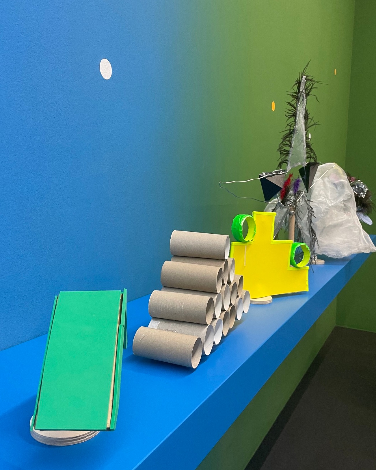 Auf einem blauen Regal vor einer blau-grün verlaufenden Wand stehen verschiedene Bastelobjekte. Links steht eine grüne Rampe auf weißen Scheiben, daneben eine Pyramide aus braunen Pappröhren. Rechts befindet sich ein gelbes Pappmodell mit grünen Ringen sowie eine Dekoration aus grünem Material und Plastikfolie. (KI-Alternativtext)