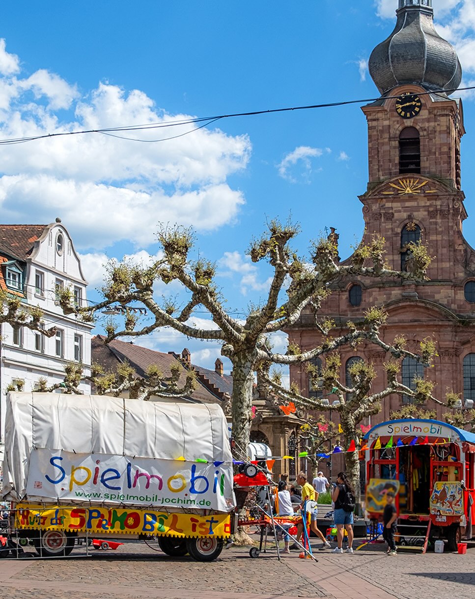 Dieser Alternativtext wurde KI-generiert: Ein farbenfrohes Stadtbild mit einem Kirchturm und einem Platz. Im Vordergrund befindet sich ein Marktstand mit Spielzeug und Menschen.