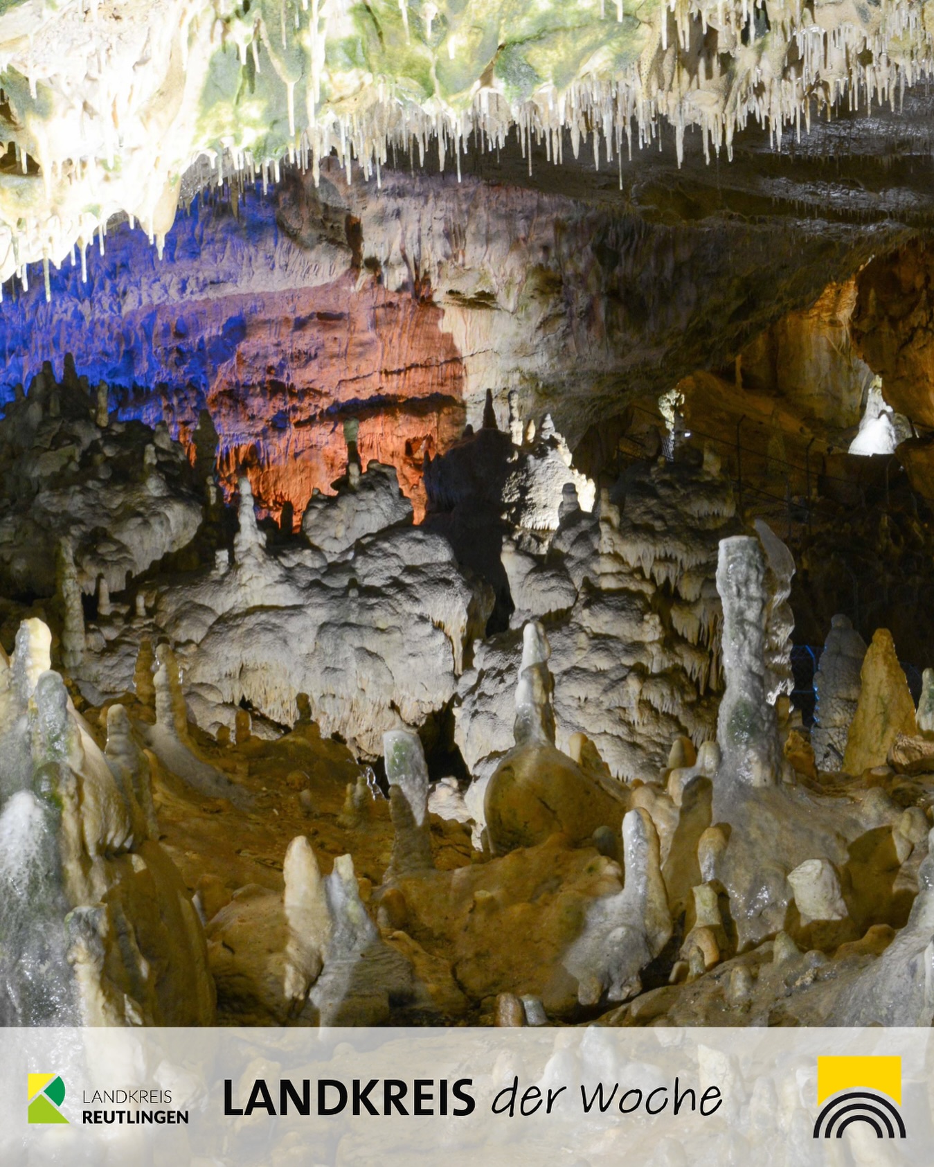 Das Bild zeigt das Innere einer Höhle mit Stalaktiten und Stalagmiten. Der Fels ist in verschiedenen Farben beleuchtet.