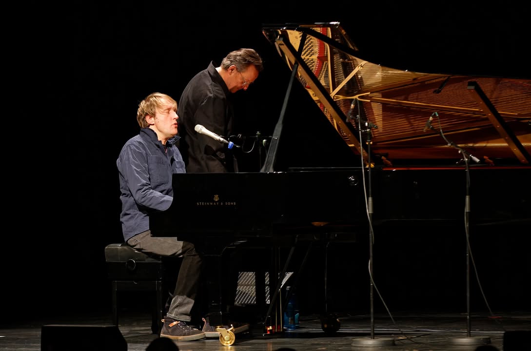 Dieser Alternativtext wurde KI-generiert: Zwei Personen sitzen vor einem schwarzen Flügel auf einer Bühne. Das Bild ist in Schwarzweiß gehalten und zeigt eine darstellende Kunstsituation.