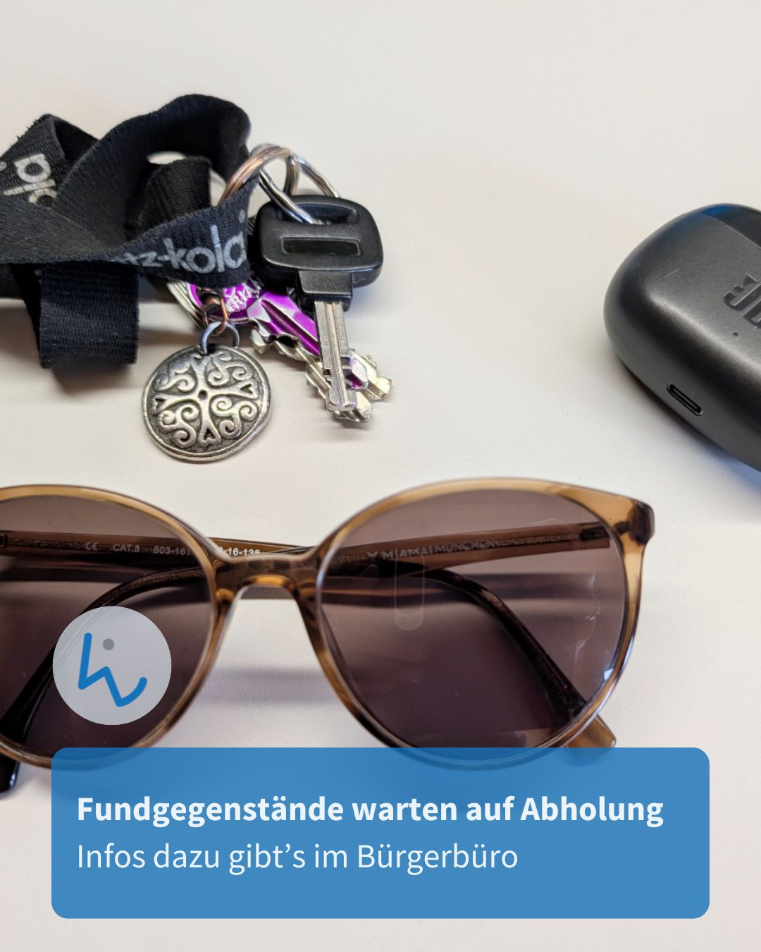 Ein Schlüsselbund mit Lanyard, eine Sonnenbrille und ein schwarzes Etui liegen auf einer hellen Fläche. Ein blauer Textkasten unten lautet: „Fundgegenstände warten auf Abholung. Infos dazu gibt’s im Bürgerbüro“. (KI-Alternativtext)