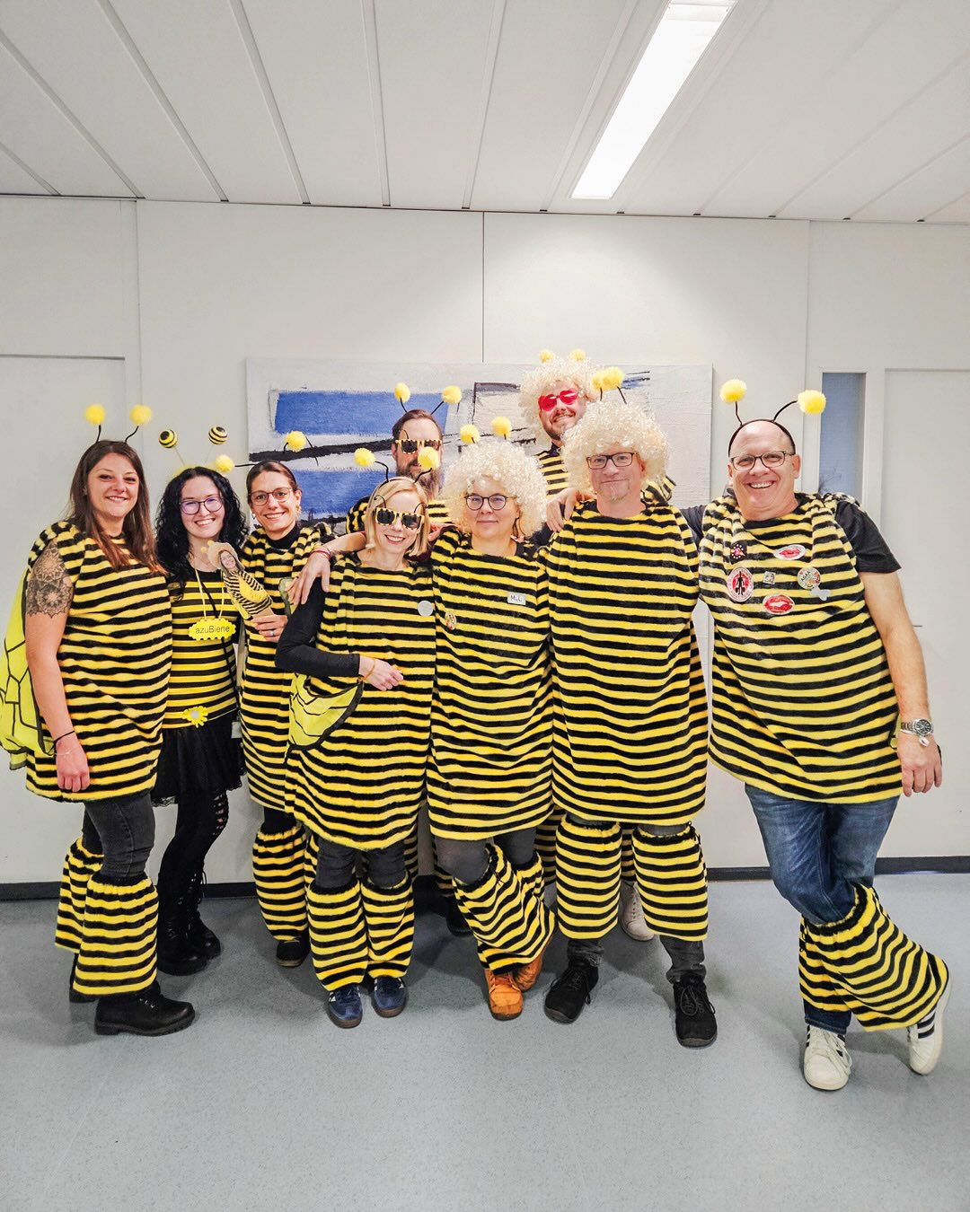 Dieser Alternativtext wurde KI-generiert: Eine Gruppe von Menschen steht lächelnd vor einer hellen Wand. Sie tragen überwiegend schwarz-gelb gestreifte Kostüme mit Antennen und Flügeln.