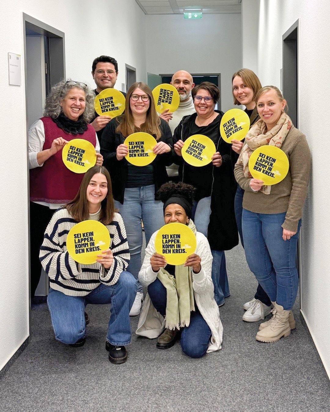 Dieser Alternativtext wurde KI-generiert: Eine Gruppe von Personen steht lächelnd in einem Innenraum zusammen. Sie halten Schilder mit Aufschriften in der Hand.
