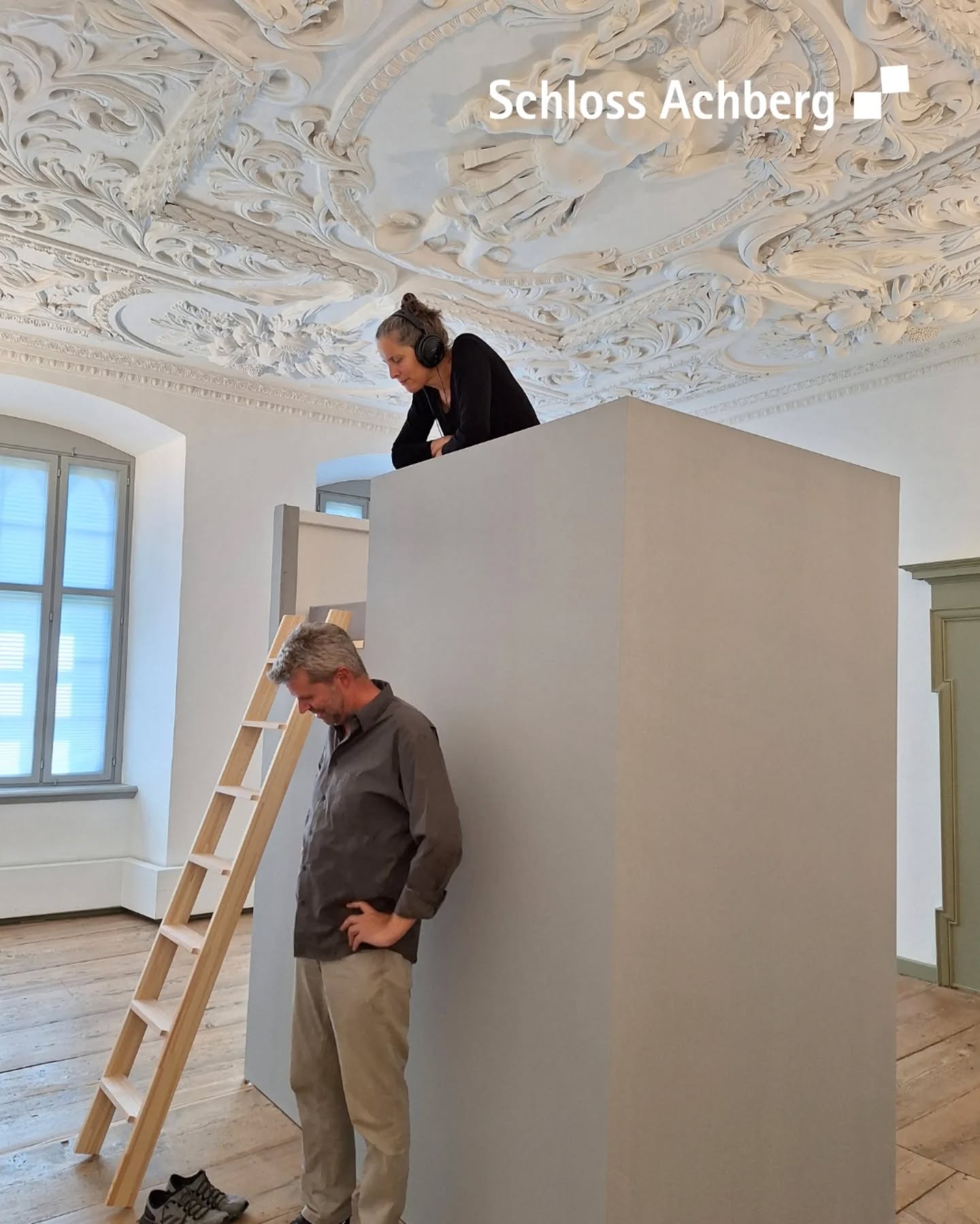 Ein Innenraum mit einer Person auf einer erhöhten Struktur und einer anderen Person, die neben einer Leiter steht. Die Decke ist mit Stuck verziert.