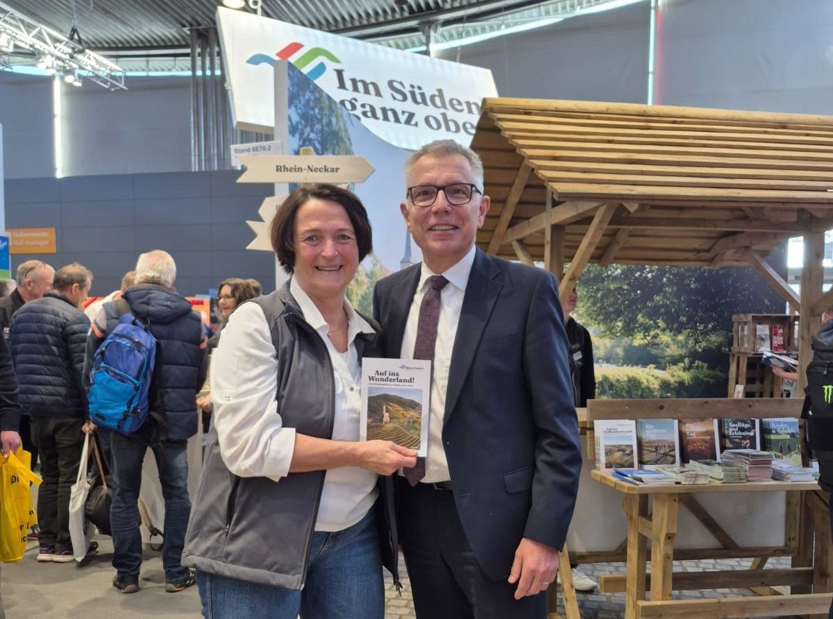 Dieser Alternativtext wurde KI-generiert: Zwei Personen stehen vor einem Stand mit Werbematerialien und einer Landschaftsdarstellung. Im Hintergrund sind weitere Menschen auf einer Veranstaltung zu sehen.