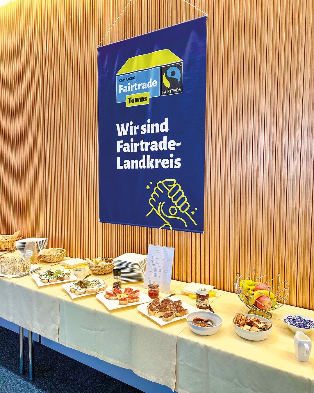 Dieser Alternativtext wurde KI-generiert: Ein blaues Banner mit Aufschrift hängt an einer Wand über einem gedeckten Tisch mit Speisen. Im Hintergrund ist ein Buffet mit verschiedenen Häppchen und Obst zu sehen.