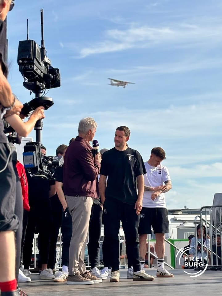 Ein Mann mit Mikrofon interviewt einen anderen Mann auf einer Bühne vor Kameras, im Hintergrund fliegt ein Flugzeug am blauen Himmel. Ein dritter Mann steht daneben und blickt auf sein Handy. (KI-Alternativtext)