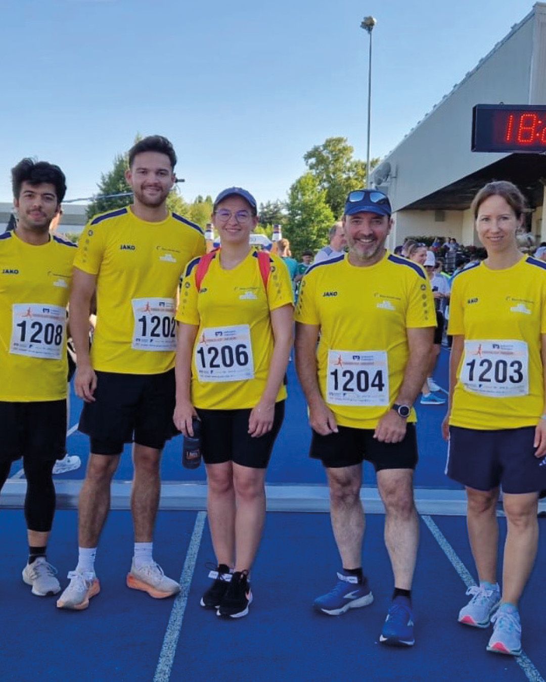 Fünf Personen in gelben T-Shirts und dunklen Shorts stehen nebeneinander auf einer blauen Laufbahn. Sie tragen Startnummern mit den Ziffern 1208, 1202, 1206, 1204 und 1203. Im Hintergrund ist ein digitales Zeitdisplay zu sehen. (KI-Alternativtext)