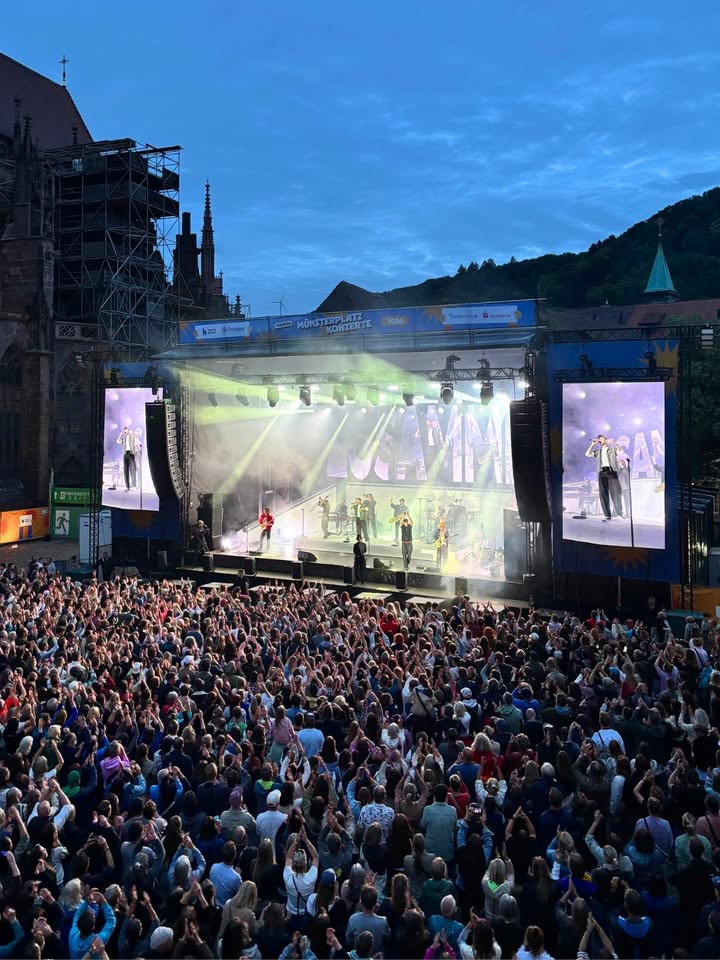 Große Menschenmenge vor einer beleuchteten Bühne bei Dämmerung, auf der Musiker auftreten; große Bildschirme zeigen die Künstler, im Hintergrund historische Gebäude und Berge. (KI-Alternativtext)