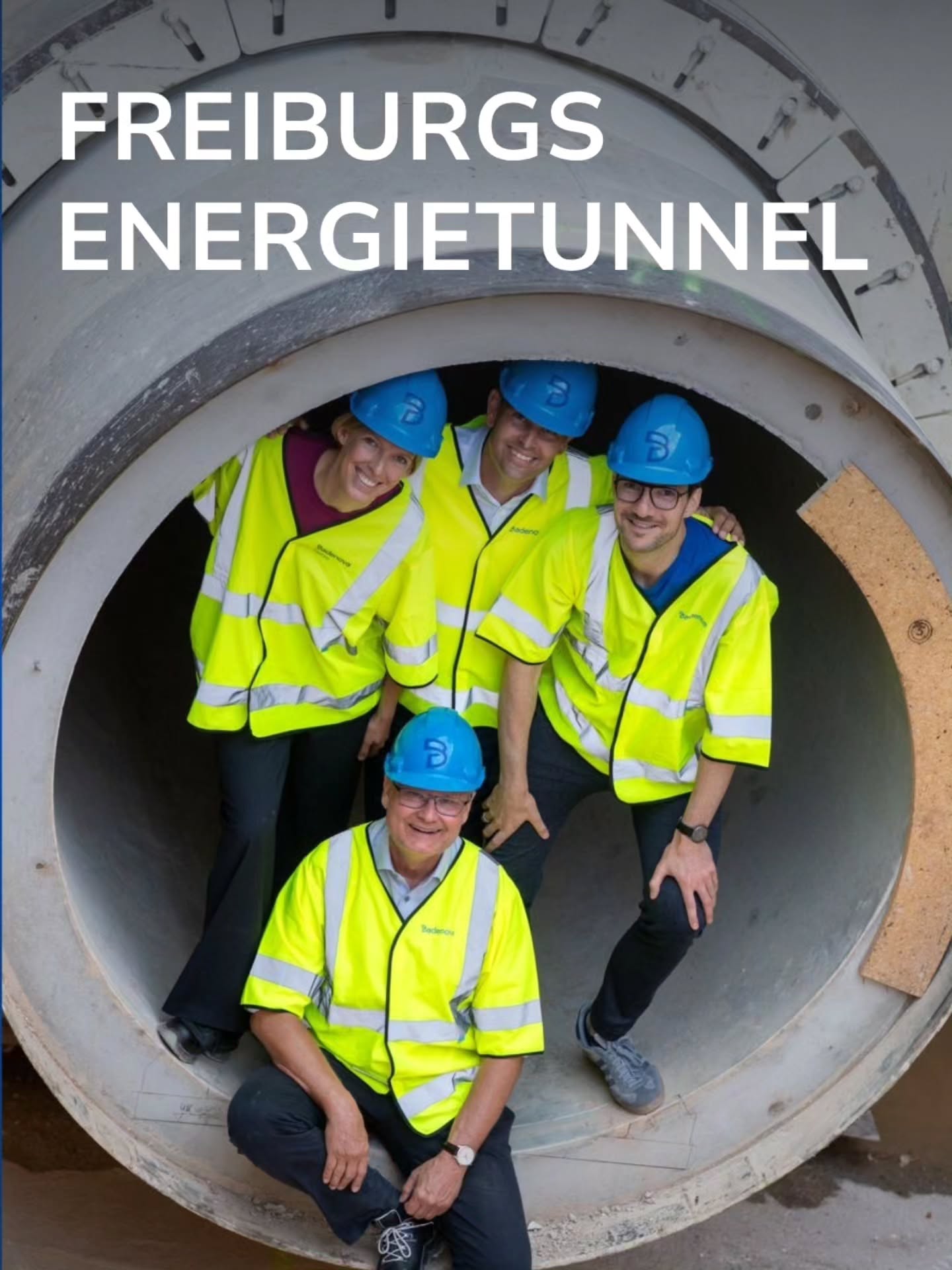 Vier Personen in blauen Schutzhelmen und gelben Warnwesten posieren lächelnd in einem großen Betonrohr. Oben ist der weiße Schriftzug „FREIBURGS ENERGIETUNNEL“ zu sehen. (KI-Alternativtext)