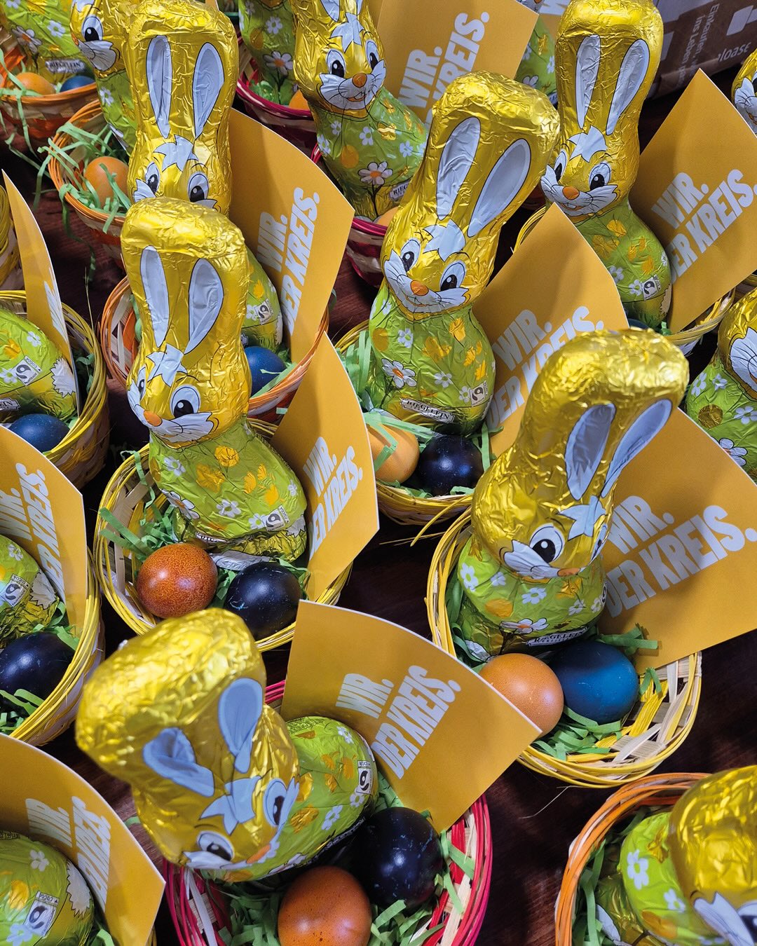 Dieser Alternativtext wurde KI-generiert: Ein hoher Stapel von Osterhasen aus Schokolade in bunten Folien steht in kleinen Körbchen. Im Hintergrund sind weitere Ostereier und Körbe zu sehen.