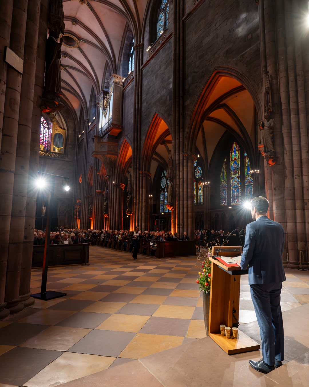Dieser Alternativtext wurde KI-generiert: Innenraum einer beeindruckenden Kirche mit hohen Gewölben und Buntglasfenstern. Eine Person steht mit dem Rücken zum Betrachter an einem Pult und spricht vor einer Menschengruppe.