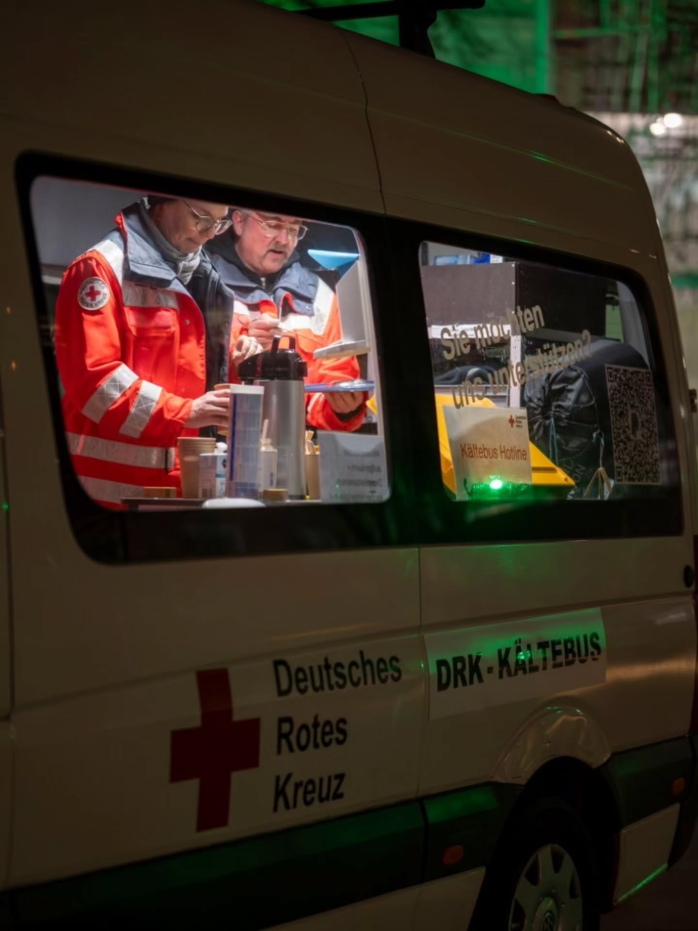 Dieser Alternativtext wurde KI-generiert: Ein weißer Transporter mit dem Logo des Deutschen Roten Kreuzes steht im Dunkeln. Durch das Fenster ist eine Person mit einer roten Weste zu sehen, die an einem Tisch arbeitet.