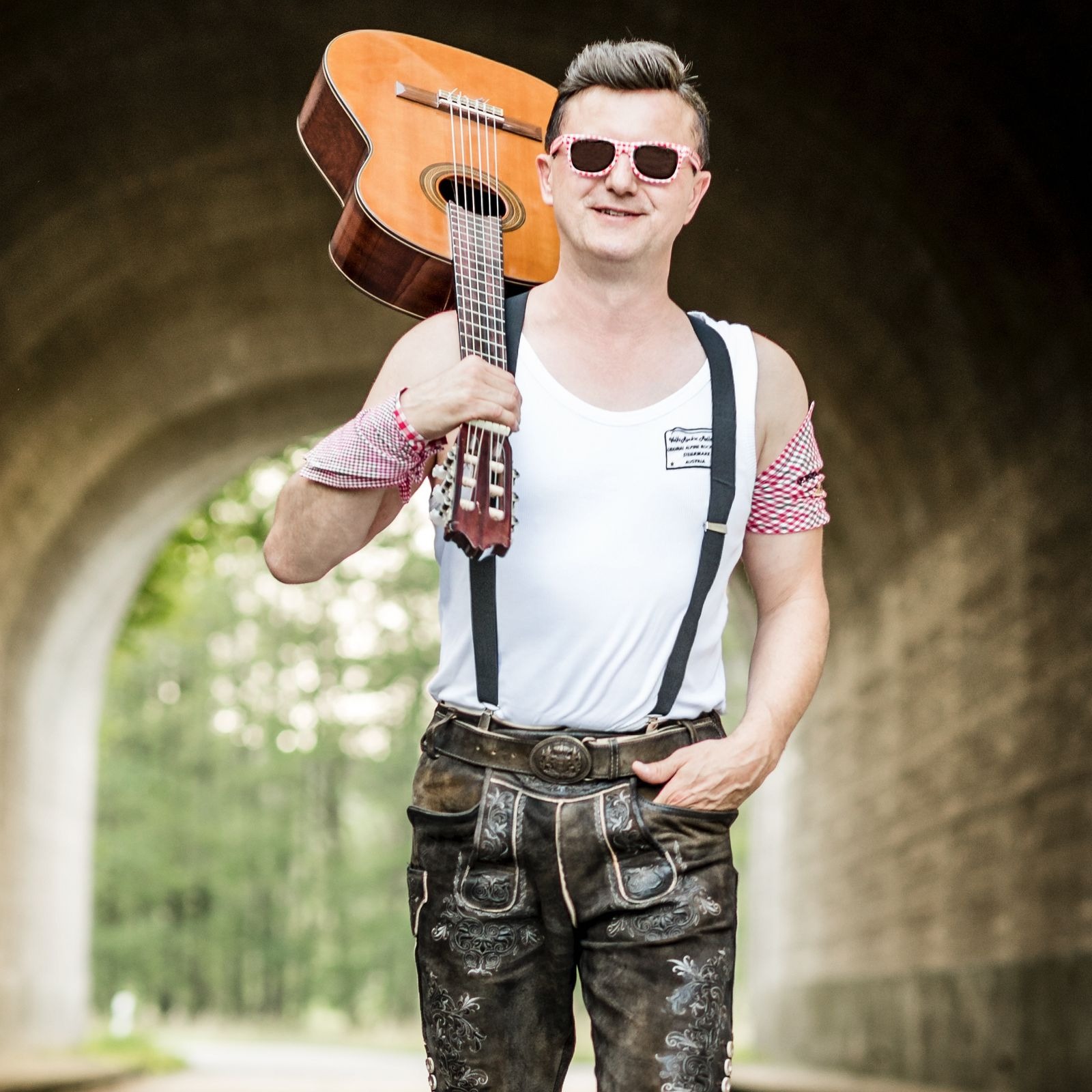 Eine Person steht vor einem Tunnelbogen und hält eine akustische Gitarre über der Schulter. Die Person trägt eine weiße Weste mit Logo, Hosenträger, dunkle bestickte Lederhosen, rote karierte Ärmel und eine rosa gemusterte Sonnenbrille. (KI-Alternativtext)