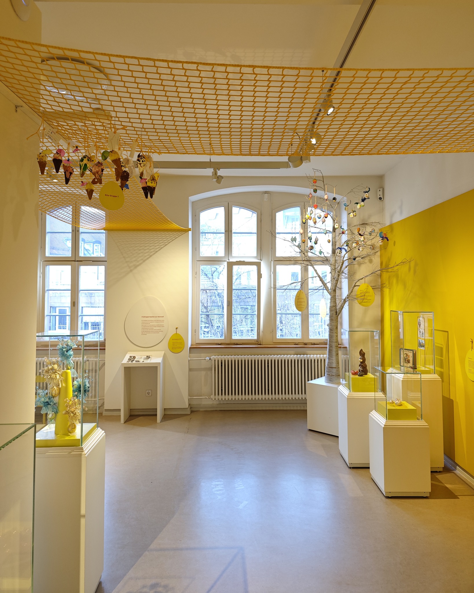 Dieser Alternativtext wurde KI-generiert: Ein heller Raum mit gelben Akzenten und einer Installation, die an Äste mit Ornamenten erinnert. Im Hintergrund sind Fenster und Vitrinen mit Ausstellungsstücken zu sehen.