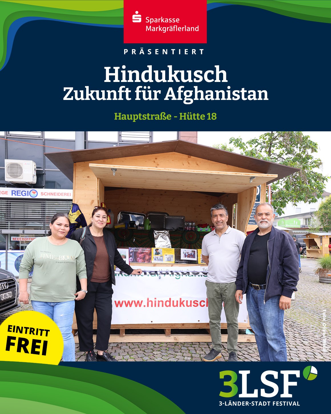 Vier Personen stehen vor einem Holzstand mit der Aufschrift „Hindukusch – Zukunft für Afghanistan“ und dem Logo der Sparkasse Markgräflerland. Ein gelber Kreis mit „Eintritt frei“ und das Logo „3LSF – 3-Länder-Stadt Festival“ sind ebenfalls sichtbar. Der Stand befindet sich auf einer gepflasterten Straße, im Hintergrund sind Bäume und ein Gebäude zu erkennen. (KI-Alternativtext)