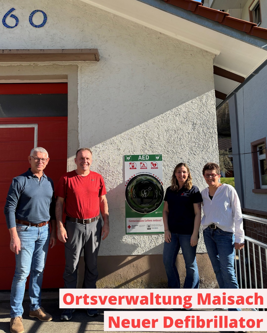 Vier Personen stehen vor einer Hauswand neben einem AED-Defibrillator in einer Wandhalterung. Über dem Eingang ist die Hausnummer 60 angebracht. Ein Textfeld unten besagt: „Ortsverwaltung Maisach – Neuer Defibrillator“. (KI-Alternativtext)