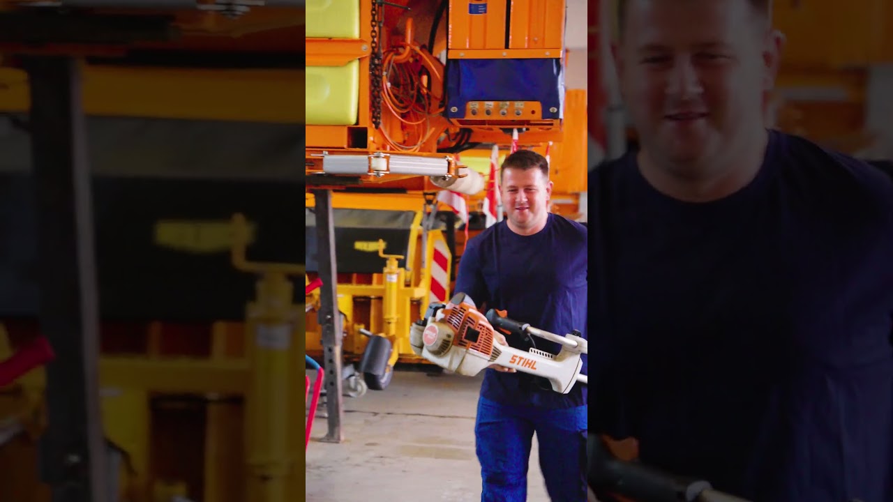 Dieser Alternativtext wurde KI-generiert: Eine Person hält eine orangefarbene Maschine mit einem Griff. Im Hintergrund sind verschiedene gelbe und graue Geräte zu sehen.