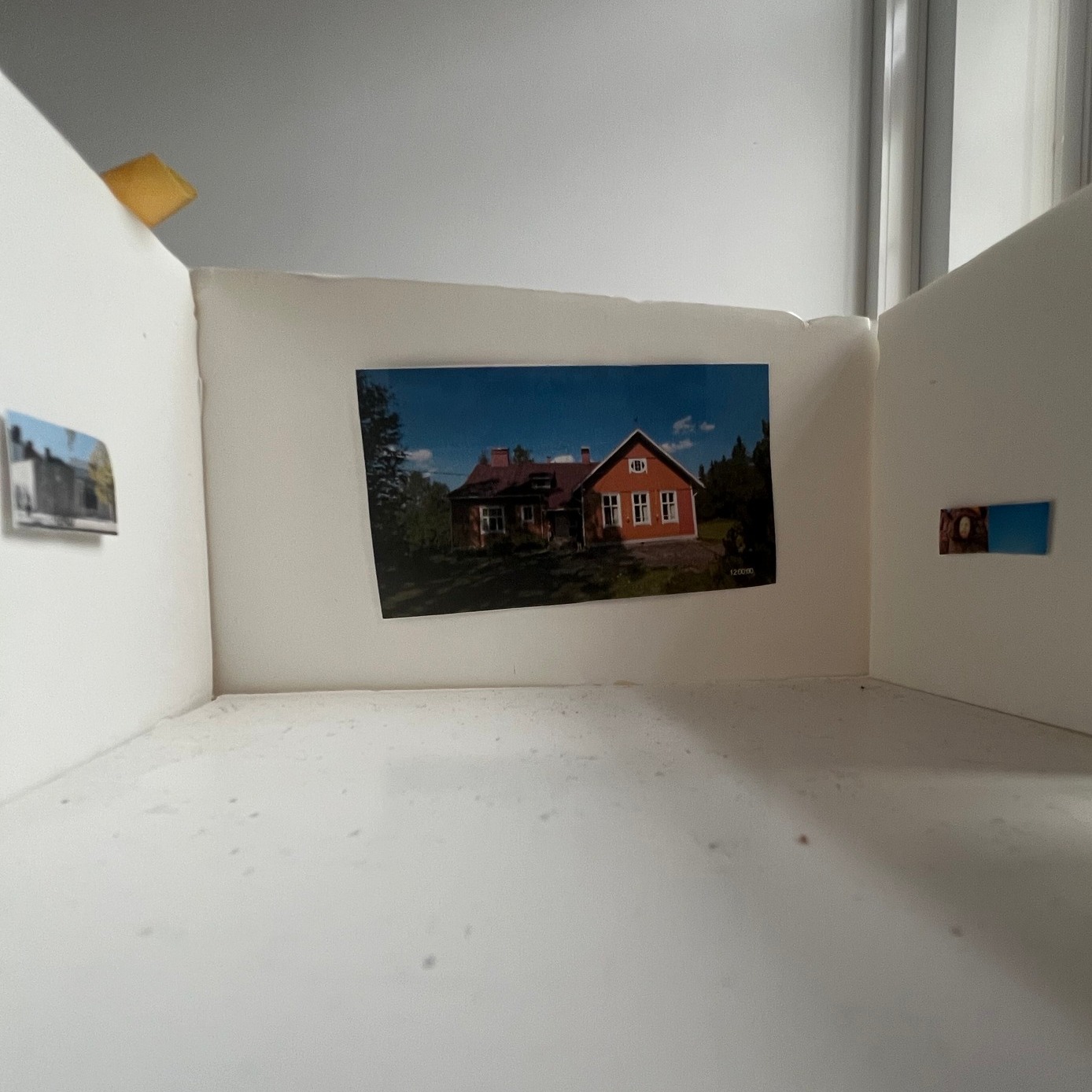 Dieser Alternativtext wurde KI-generiert: Eine Ansicht aus einem Fensterrahmen, der eine weitere Abbildung von einem Haus zeigt. Die Aufnahme ist in einer hellen Umgebung entstanden.