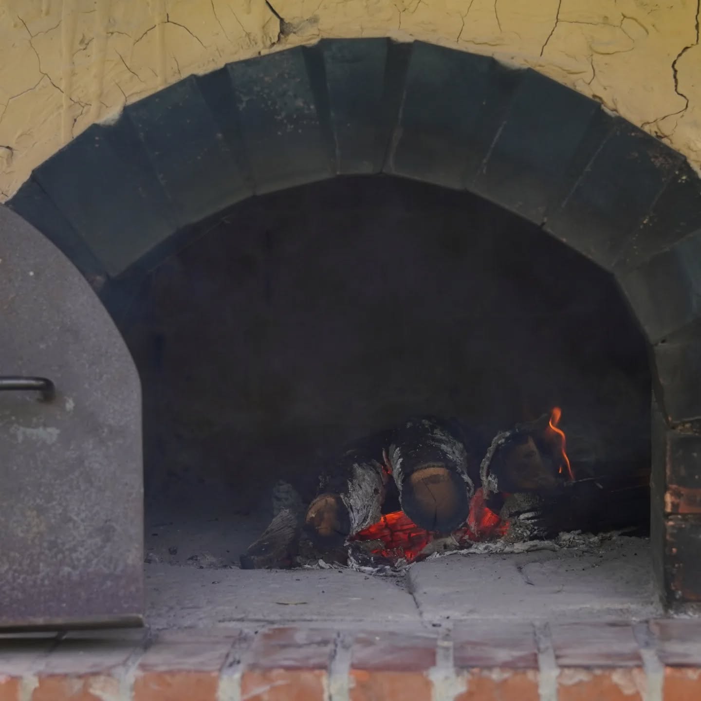 Dieser Alternativtext wurde KI-generiert: Ein alter, gemauerter Ofen mit einem dunklen Innenraum. Im Inneren lodern Flammen und es liegt Holz.