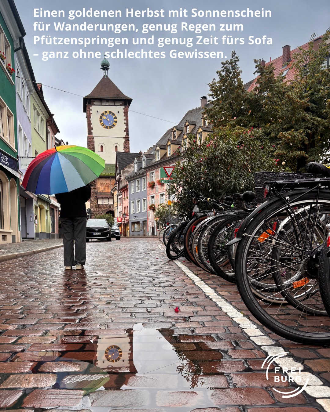 Dieser Alternativtext wurde KI-generiert: Eine Straßenszene mit einem Gebäude im Hintergrund und regennassen Pflastersteinen. Fahrräder und eine Person mit Regenschirm sind im Vordergrund zu sehen.
