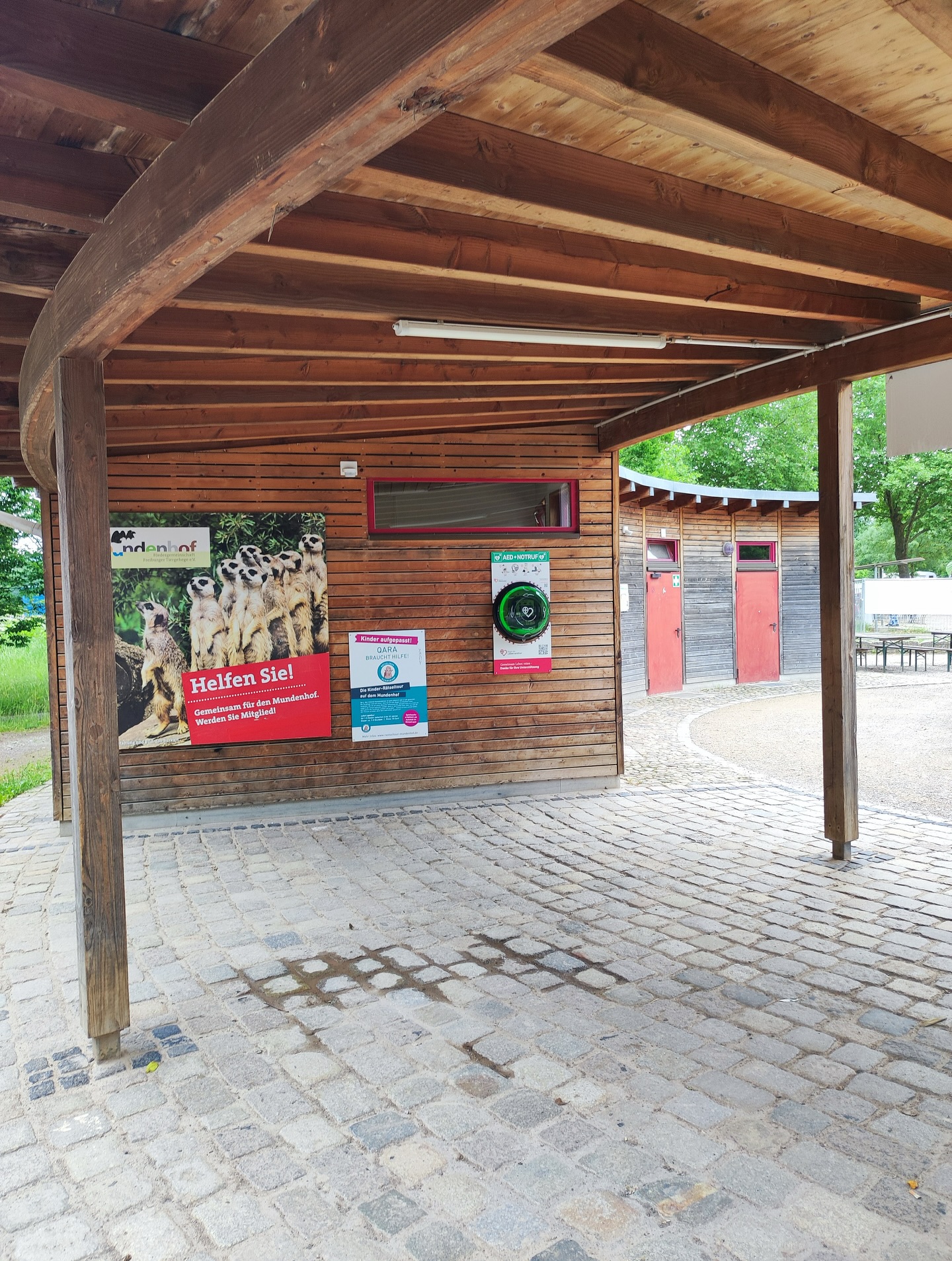 Das Foto zeigt ein Gebäude mit Holzwänden unter einem Holzdach. An der Wand hängen mehrere Schilder.