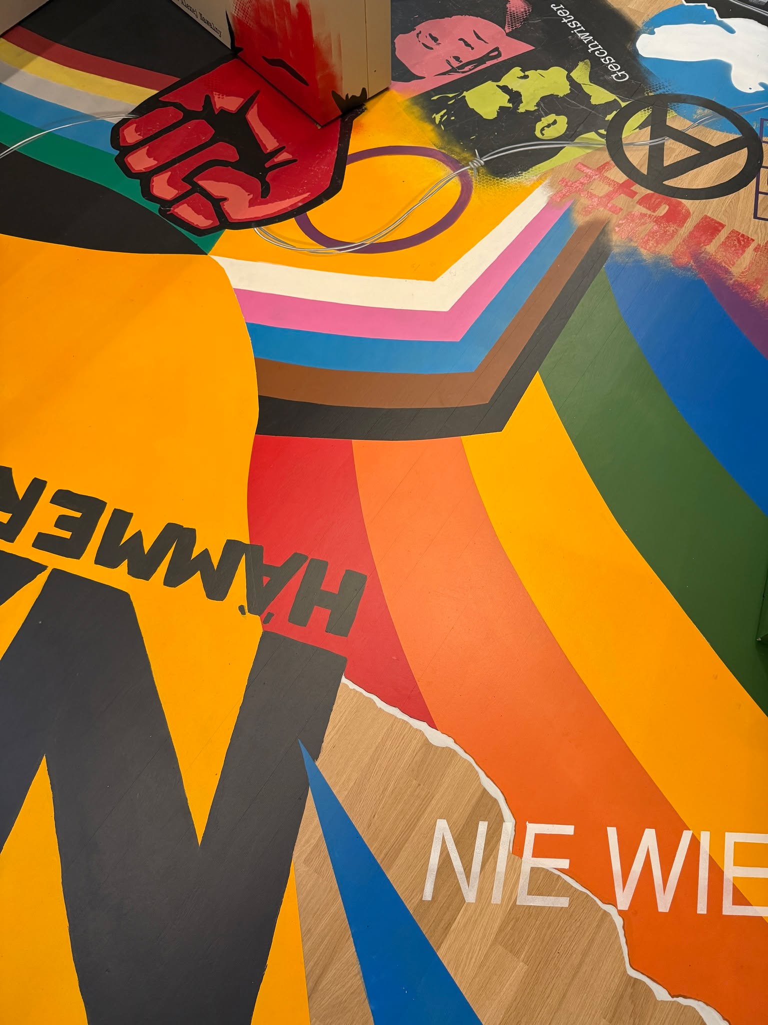 Bunte Wandgestaltung mit stilisierter roter Faust, Regenbogenfarben, dem Text „HÄMMER“ und „NIE WIE“ sowie einem Anarchie-Symbol und Gesichtern im Hintergrund. (KI-Alternativtext)