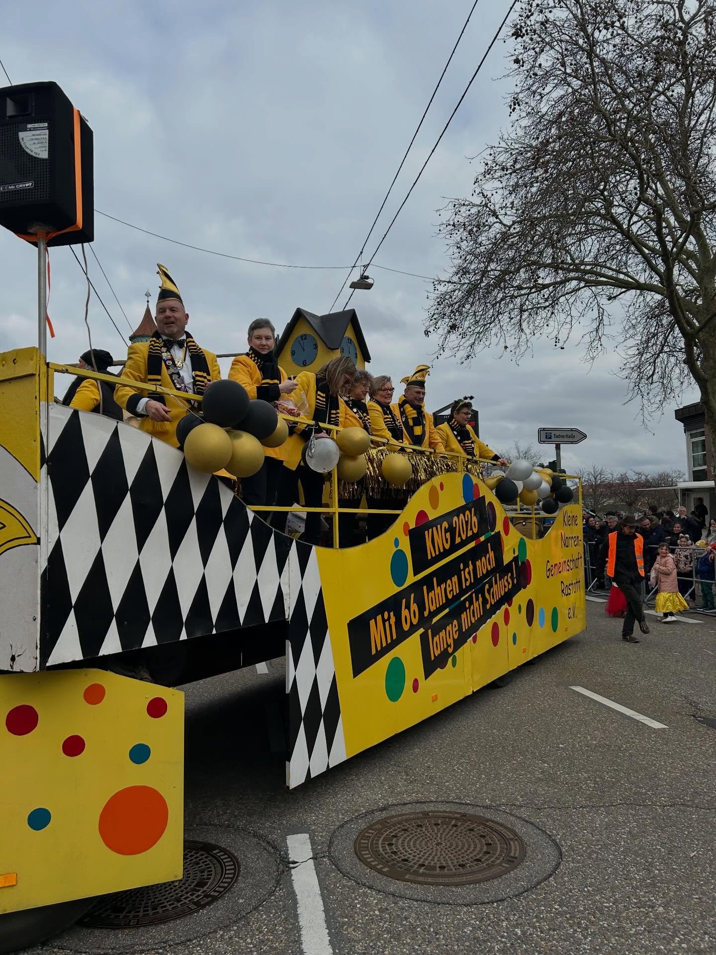 Dieser Alternativtext wurde KI-generiert: Ein farbenfroher Festwagen fährt eine Straße entlang. Der Wagen ist mit bunten Punkten und Schildern dekoriert und wird von Menschen in auffälligen Kostümen begleitet.