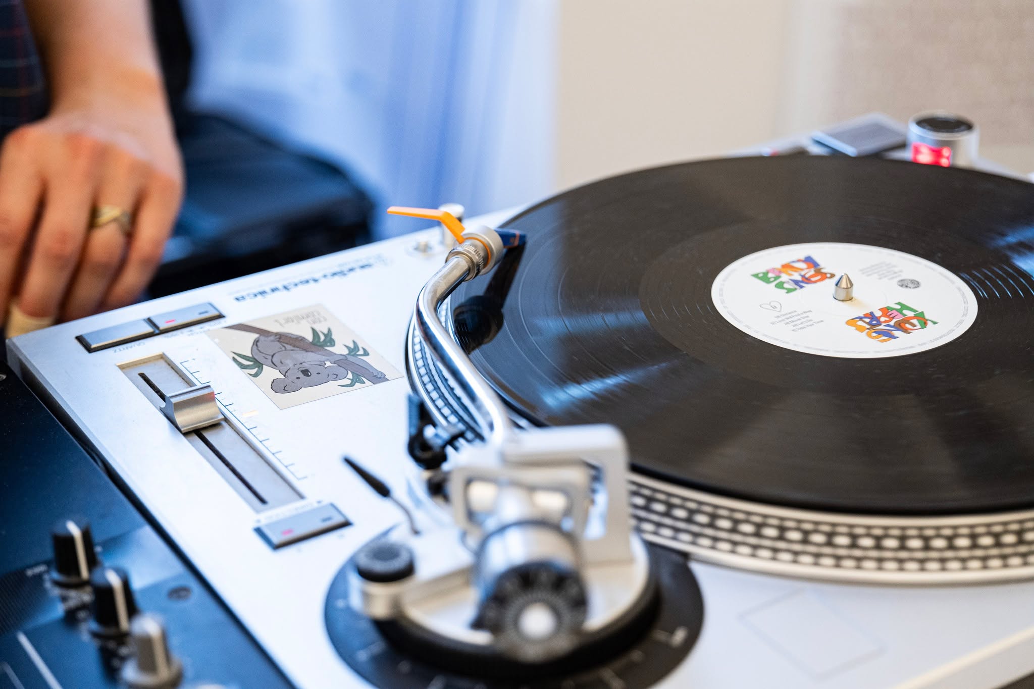 Ein weißer Plattenspieler mit einer schwarzen Schallplatte, auf deren Label "BODY SONG" und "LOVE HEAL" steht. Der Tonarm liegt auf der Platte, während im Hintergrund eine unscharfe Hand die Bedienelemente bedient. Auf dem Gerät befindet sich ein Aufkleber mit einem Koala. (KI-Alternativtext)