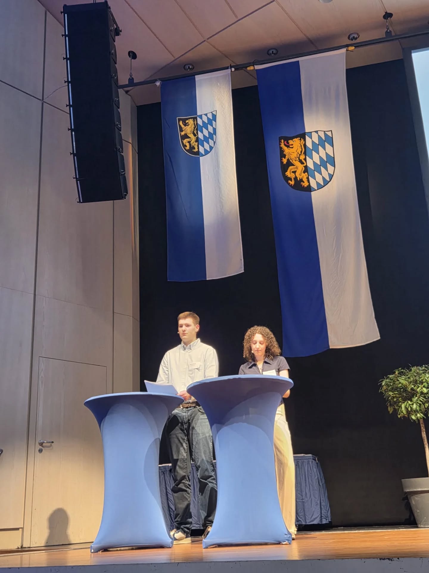 Dieser Alternativtext wurde KI-generiert: Zwei Personen stehen vor einem Pult auf einer Bühne. Im Hintergrund hängen Flaggen mit Wappen an einer dunklen Wand.
