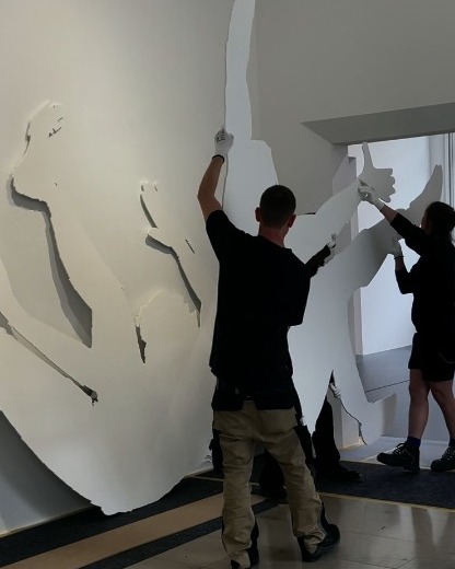 Dieser Alternativtext wurde KI-generiert: Zwei Personen tragen weiße Handschuhe und halten ausgeschnittene Formen an einer hellen Wand fest. Das Bild ist farbig und zeigt eine Innenraumansicht.