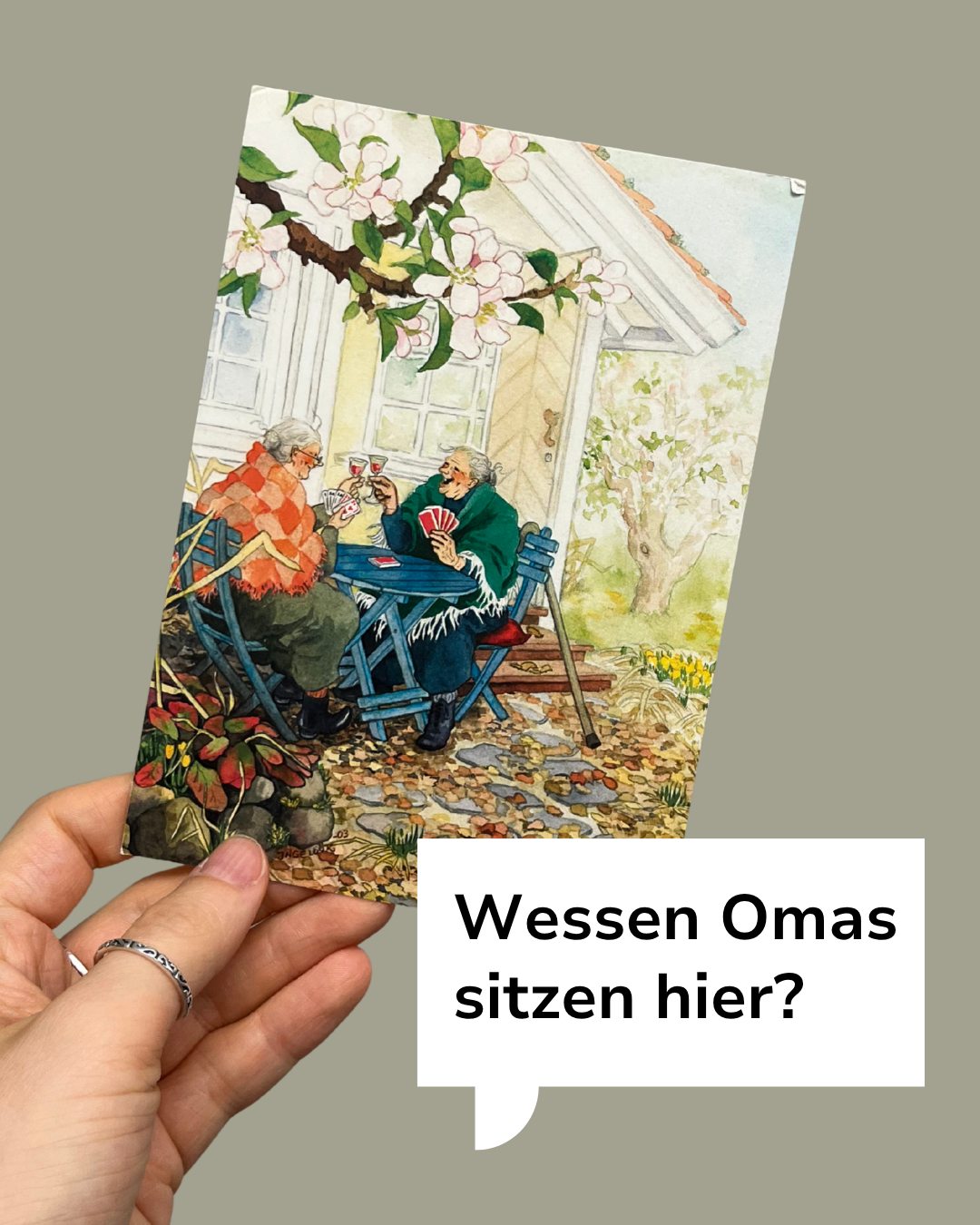 Eine Hand hält eine Postkarte, auf der zwei ältere Personen an einem blauen Tisch im Freien Karten spielen und Wein trinken. Die Szene ist von blühenden Bäumen und Pflanzen umgeben. Ein Textfeld fragt: „Wessen Omas sitzen hier?“ (KI-Alternativtext)