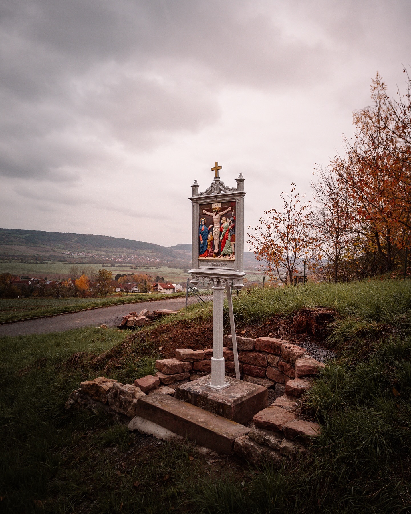 Ein Bild zeigt ein kleines, weißes Denkmal mit einem religiösen Bild in einer ländlichen Gegend. Im Hintergrund ist eine hügelige Landschaft mit Bäumen zu sehen.